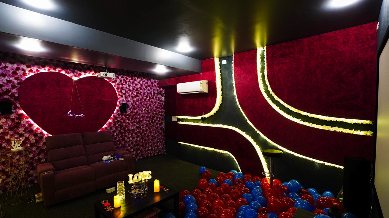 Romantic private theatre room with red heart wall décor, cozy sofa seating, candles on table, and colorful balloons creating an intimate celebration setup.